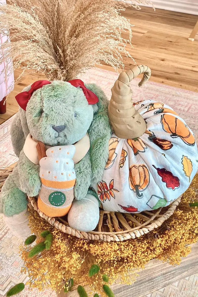 Baby shower presents featuring a plush green bunny with red bows, a fabric pumpkin wrapped in autumn-themed swaddle, and a pumpkin spice latte baby sock set