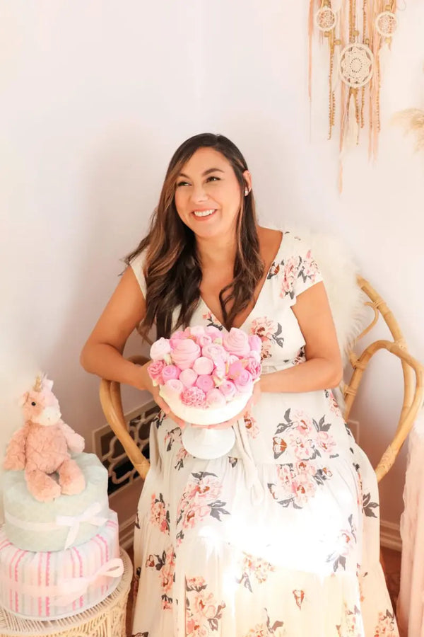 Gifts for baby showers with a smiling woman holding a pink diaper bouquet and a diaper cake topped with a unicorn toy.